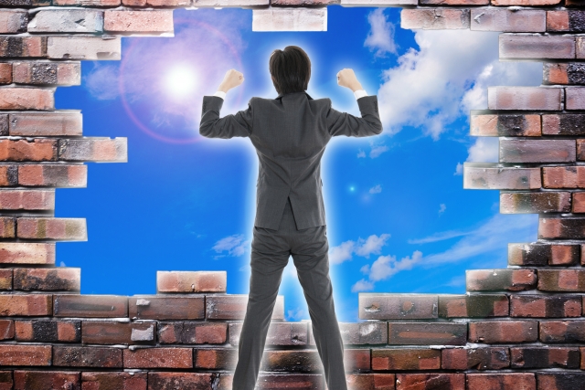 スーツ姿の男性がレンガの壁を突き破り、青空に向かって拳を上げているイメージ(営業は体育会系じゃないと無理?という不安を乗り越える象徴)