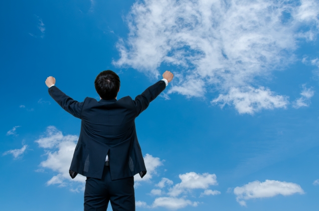 青空に向かって両手を広げるスーツ姿の男性