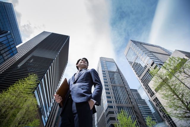 高層ビル街の中でスーツ姿の男性が立つ、営業職や転職後のキャリアアップをイメージした画像