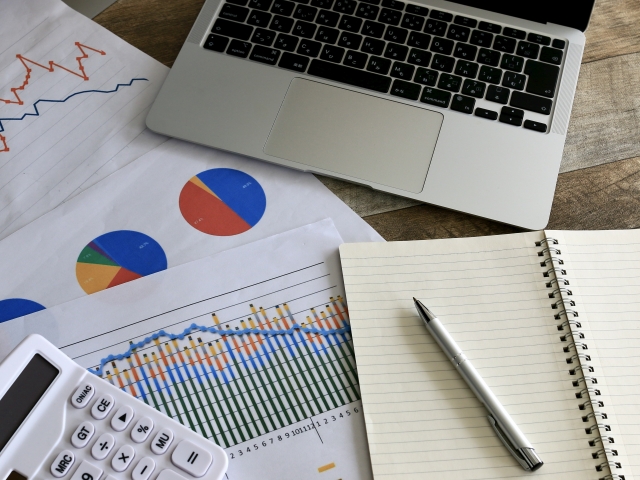 ノートパソコンの横に、グラフ資料、電卓、ノート、ペンが置かれた分析作業のイメージ画像